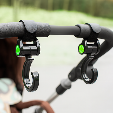 Black stroller hooks with green accents on a stroller handle against a blurred outdoor background