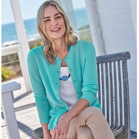 Woman sitting on a wooden bench by the sea wearing a turquoise cardigan.